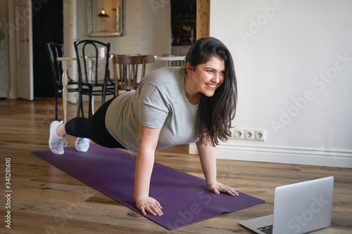 Sporty plus size woman in sportswear working out at home, doing plank on yoga mat in front of open laptop, repeating instructions by professional fitness trainer watching online video tutorial