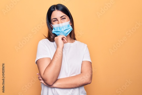 Young woman wearing protection mask for coronavirus disease over yellow background smiling looking confident at the camera with crossed arms and hand on chin. Thinking positive. © Krakenimages.com