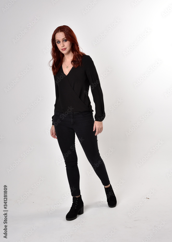Portrait of a pretty girl with red hair wearing black jeans, boots and a blouse. full length standing pose, facing the camera, on a studio background. Portrait of a pretty girl with red hair wearing black jeans, boots and a blouse. full length standing pose, facing the camera, on a studio background.