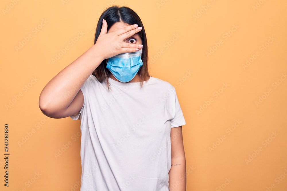 Young woman wearing protection mask for coronavirus disease over yellow background peeking in shock covering face and eyes with hand, looking through fingers afraid