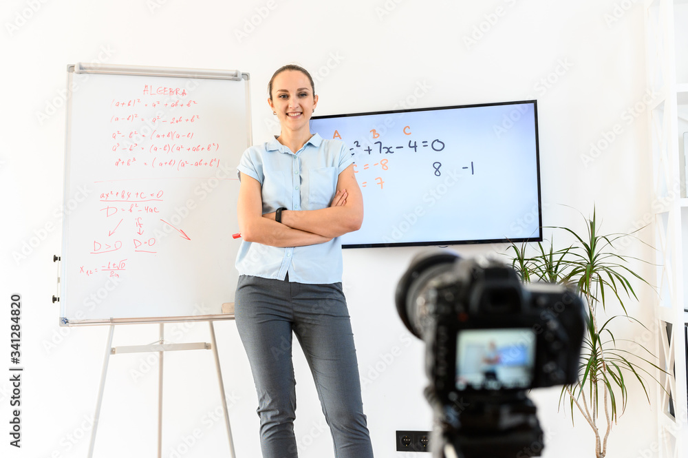 Fototapeta premium Young and confident woman teacher, tutor writes video tutorials to the camera, uses a flip chart and monitor for classes. She is standing with arms crossed and looking at the camera.