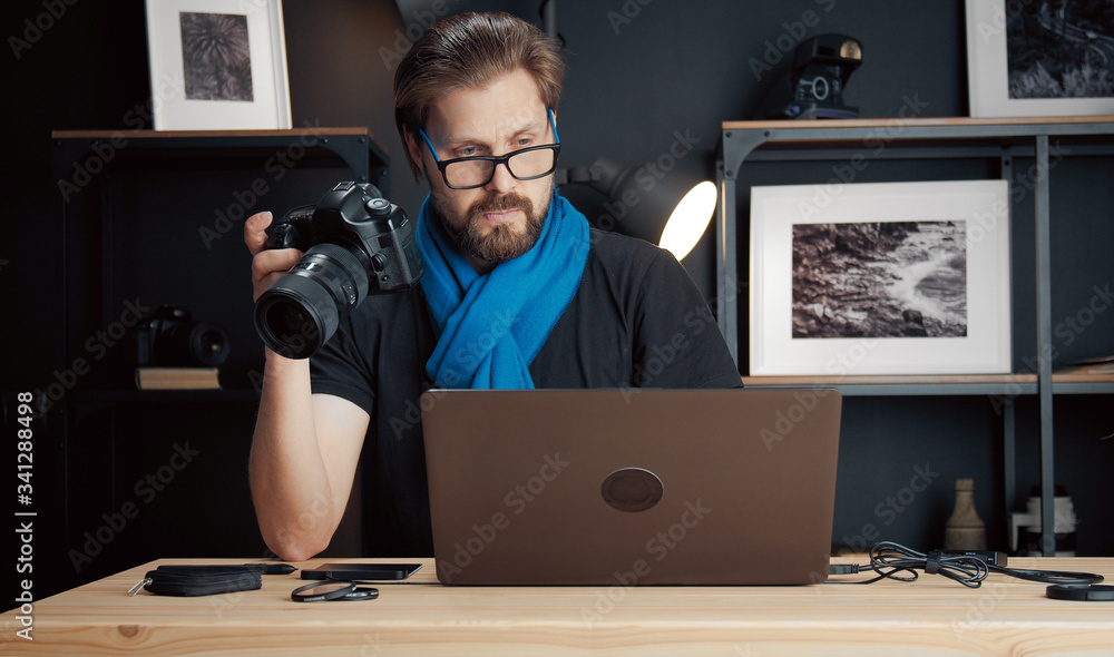 Professional photographer holding camera in one hand while looking at laptop screen sitting at desk Professional photographer holding camera in one hand while looking at laptop screen sitting at desk