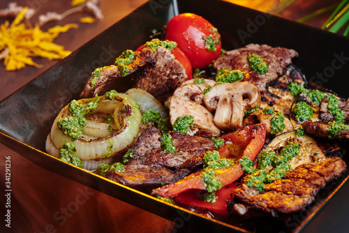 Grilled vegetables: mushrooms, bell pepper, eggplant with grilled meat  in a black square matte plate among greens on a gray smoky background with warm lighting. Space. Close up