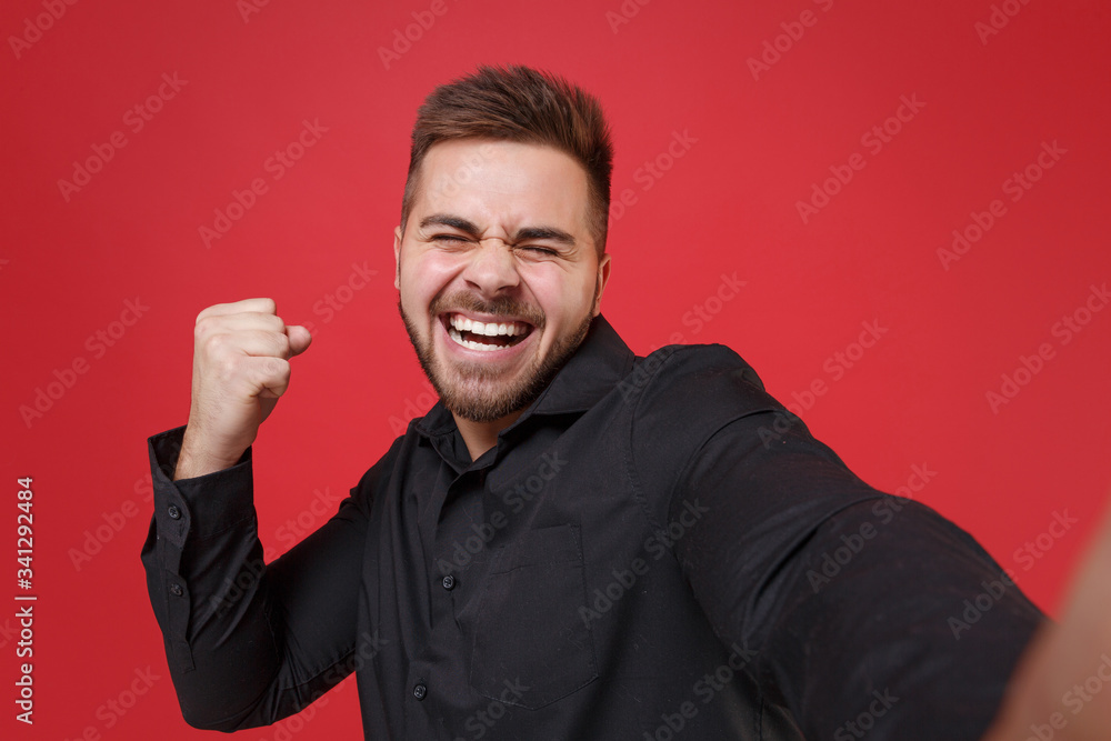 Close up of happy young bearded guy 20s in classic black shirt posing isolated on red background. People lifestyle concept. Mock up copy space. Doing selfie shot on mobile phone, doing winner gesture.