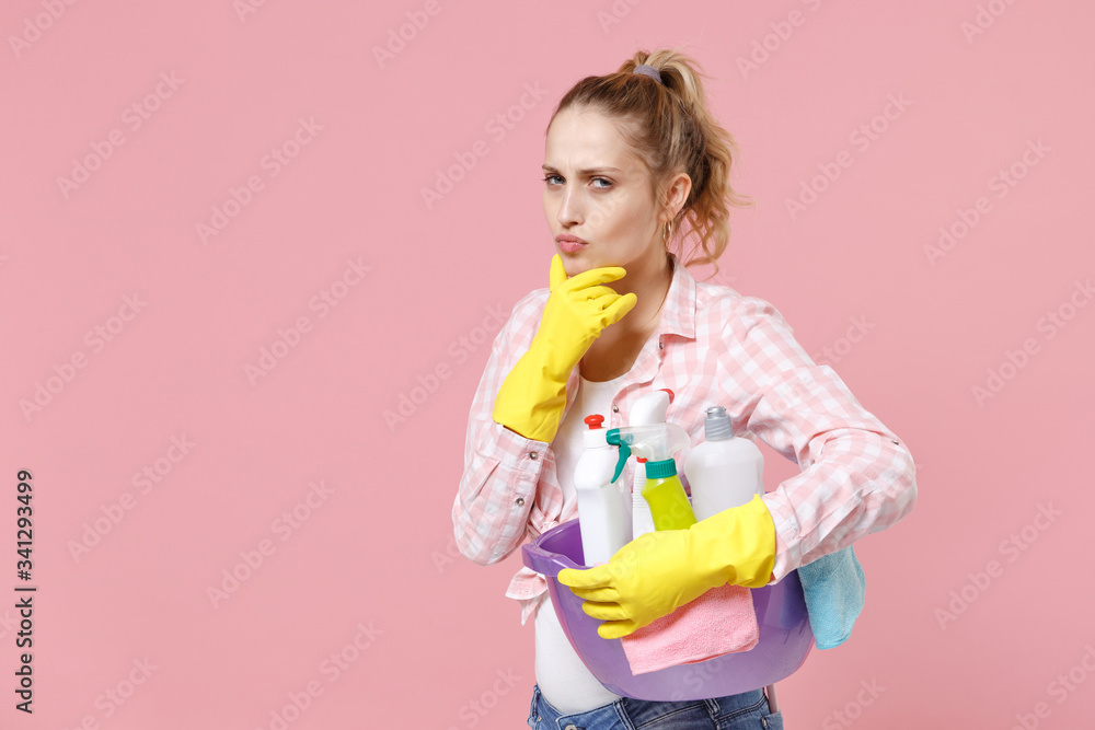 Pensive young woman housewife in rubber gloves hold basin with detergent bottles washing cleansers doing housework isolated on pink background studio. Housekeeping concept. Put hand prop up on chin. Pensive young woman housewife in rubber gloves hold basin with detergent bottles washing cleansers doing housework isolated on pink background studio. Housekeeping concept. Put hand prop up on chin.