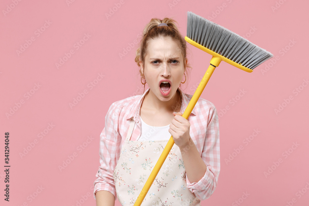 Irritated young woman housewife in casual clothes, apron doing housework isolated on pastel pink background studio portrait. Housekeeping concept. Mock up copy space. Hold in hands broom, swearing. Irritated young woman housewife in casual clothes, apron doing housework isolated on pastel pink background studio portrait. Housekeeping concept. Mock up copy space. Hold in hands broom, swearing.
