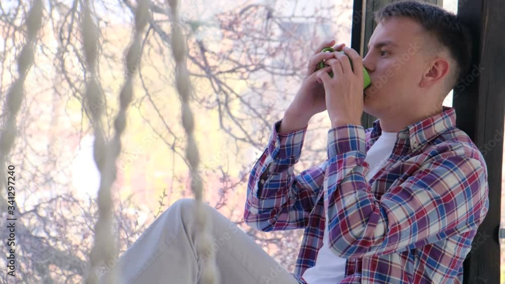 Cute young guy sitting on a window sill by the window and drinking tea at home. Home quarantine, social distancing, self isolation. Cute young guy sitting on a window sill by the window and drinking tea at home. Home quarantine, social distancing, self isolation.