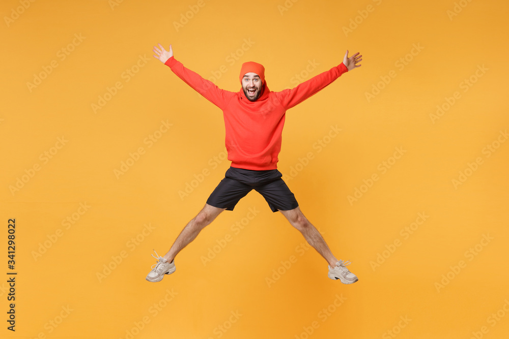 Excited young bearded fitness sporty guy 20s sportsman in hat hoodie shorts in home gym isolated on yellow background. Workout sport motivation lifestyle concept. Jumping spreading hands and legs.