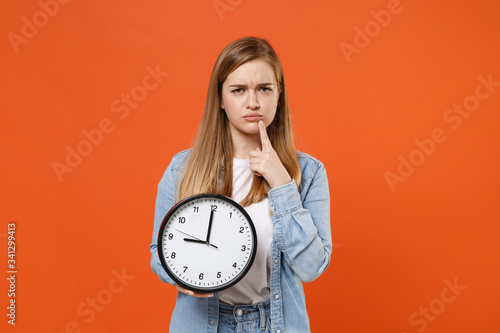 Confused pensive young woman girl in casual denim clothes isolated on orange background studio portrait. People emotions lifestyle concept. Mock up copy space. Hold clock, put hand prop up on chin.
