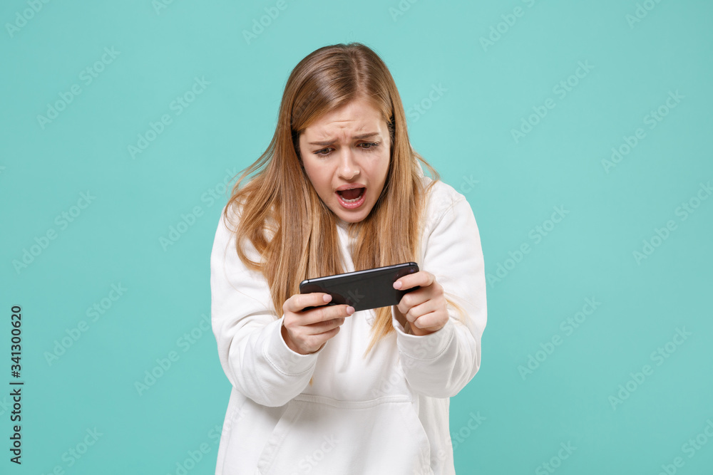 Dissatisfied young woman girl in casual white hoodie posing isolated on blue turquoise background studio portrait. People emotions lifestyle concept. Mock up copy space. Play game with mobile phone. Dissatisfied young woman girl in casual white hoodie posing isolated on blue turquoise background studio portrait. People emotions lifestyle concept. Mock up copy space. Play game with mobile phone.