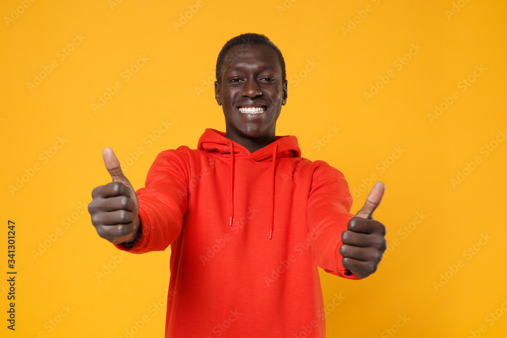 Smiling young african american man guy in red streetwear hoodie posing isolated on yellow background studio portrait. People sincere emotions, lifestyle concept. Mock up copy space. Showing thumbs up. Smiling young african american man guy in red streetwear hoodie posing isolated on yellow background studio portrait. People sincere emotions, lifestyle concept. Mock up copy space. Showing thumbs up.
