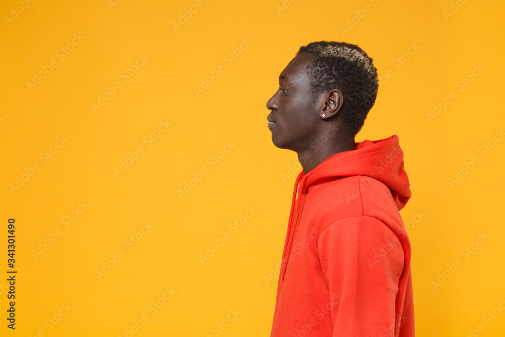 Side view of young african american man guy in red streetwear hoodie posing isolated on yellow orange background studio portrait. People emotions lifestyle concept. Mock up copy space. Looking aside. Side view of young african american man guy in red streetwear hoodie posing isolated on yellow orange background studio portrait. People emotions lifestyle concept. Mock up copy space. Looking aside.