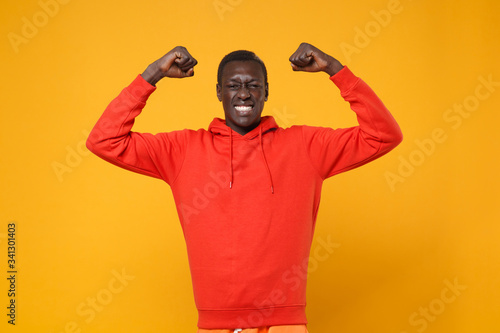 Smiling young african american man guy in red streetwear hoodie posing isolated on yellow orange wall background studio portrait. People lifestyle concept. Mock up copy space. Doing winner gesture.