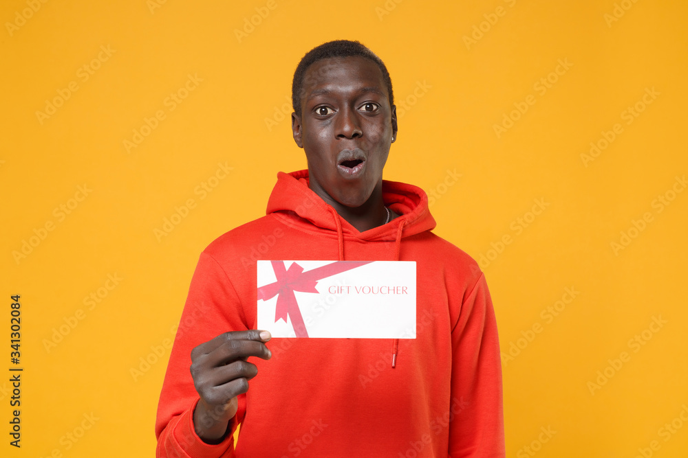 Shocked young african american man guy in red streetwear hoodie posing isolated on yellow wall background studio portrait. People emotions lifestyle concept. Mock up copy space. Hold gift certificate. Shocked young african american man guy in red streetwear hoodie posing isolated on yellow wall background studio portrait. People emotions lifestyle concept. Mock up copy space. Hold gift certificate.