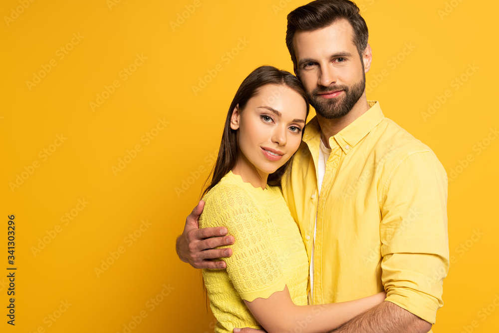 Naklejka premium Handsome man embracing beautiful smiling girlfriend and looking at camera on yellow background