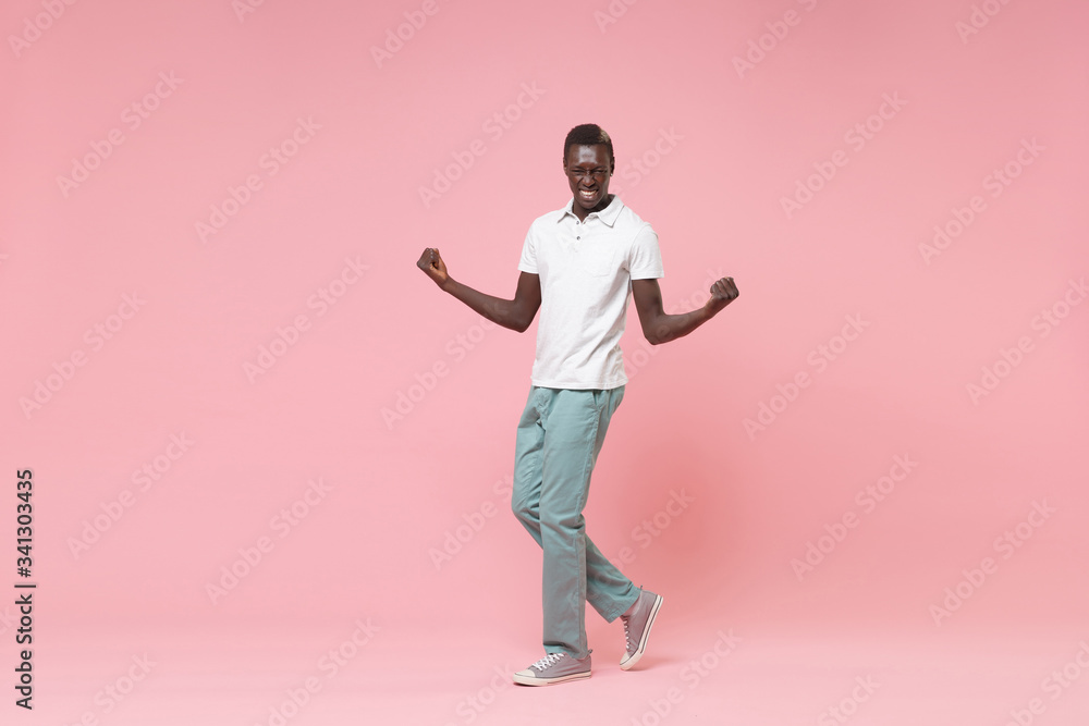 Happy young african american man guy in white polo shirt, turquoise trousers posing isolated on pastel pink wall background. People lifestyle concept. Mock up copy space. Clenching fists like winner. Happy young african american man guy in white polo shirt, turquoise trousers posing isolated on pastel pink wall background. People lifestyle concept. Mock up copy space. Clenching fists like winner.