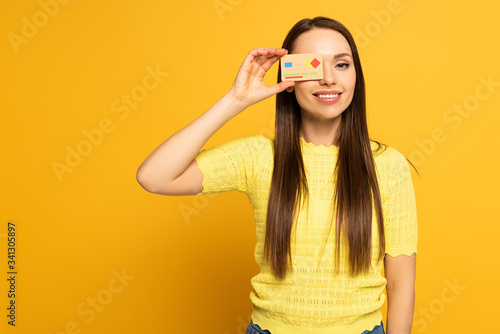 Smiling girl covering eye with model of credit card on yellow background