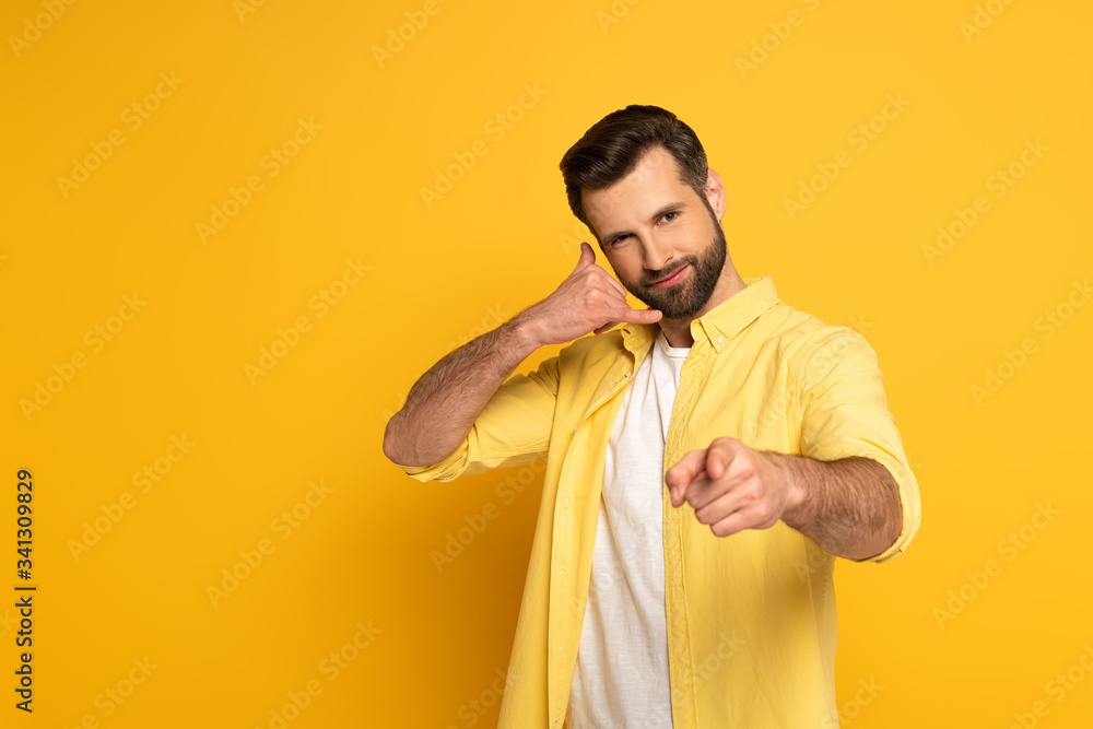 Fototapeta premium Handsome man showing call sign and pointing with finger at camera on yellow background