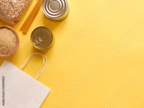 Dry goods, delivery, stockpiling, food supplies for staying home concept, donation, volunteer. Craft paper shopping bag, preserves, pasta, oatmeal, rice, on yellow background, copyspace