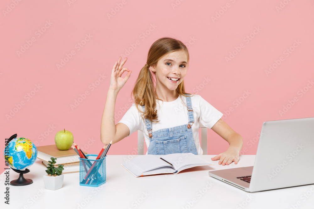 Smiling little kid schoolgirl 12-13 years old sit study at white desk with pc laptop isolated on pastel pink background. School distance education at home during quarantine concept Showing OK gesture. Smiling little kid schoolgirl 12-13 years old sit study at white desk with pc laptop isolated on pastel pink background. School distance education at home during quarantine concept Showing OK gesture.