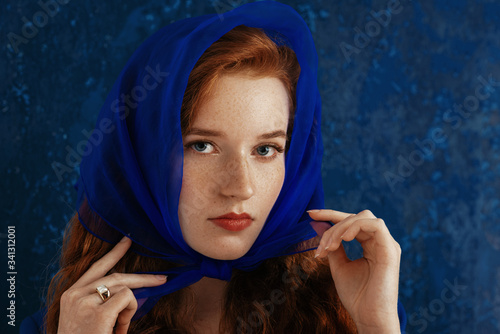 Close up fashion portrait of elegant redhead woman with freckled skin wearing luxury trendy headscarf, pearl ring, posing on dark blue background. Copy, empty space for text  photo