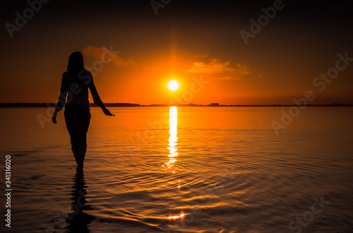 Young girl standing in the lake on sunset back ground. Happy moments. Relax for soul and body. Quiet ending of the day. Girl's silhouette on shore of the lake looks far into the distance. 
