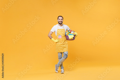 Smiling man househusband in apron rubber gloves hold basin detergent bottles washing cleansers doing housework isolated on yellow background. Housekeeping concept. Standing with arm akimbo on waist.