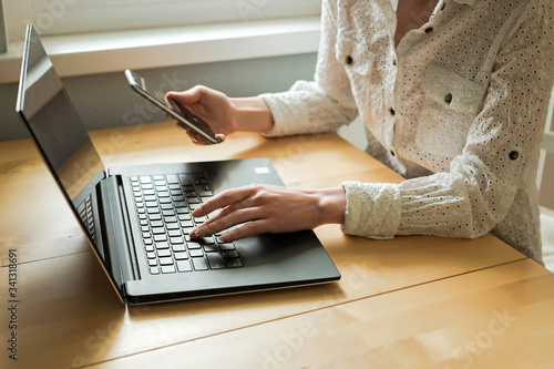 Woman's hands using laptop and smartphone on desk in home interior. Remote work. Freelance.