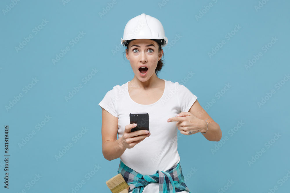 Shocked young woman in protective helmet hardhat pointing index finger on mobile phone isolated on blue background studio. Instruments accessories for renovation apartment room. Repair home concept. Shocked young woman in protective helmet hardhat pointing index finger on mobile phone isolated on blue background studio. Instruments accessories for renovation apartment room. Repair home concept.