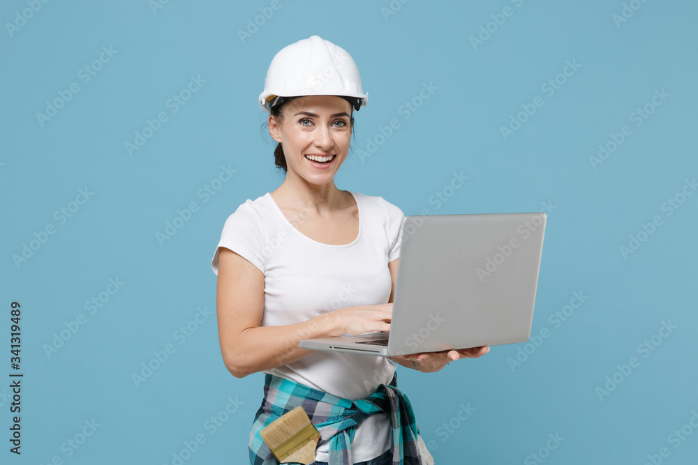 Cheerful young woman in protective helmet hardhat working on laptop pc computer isolated on pastel blue background studio. Instruments accessories for renovation apartment room. Repair home concept. Cheerful young woman in protective helmet hardhat working on laptop pc computer isolated on pastel blue background studio. Instruments accessories for renovation apartment room. Repair home concept.