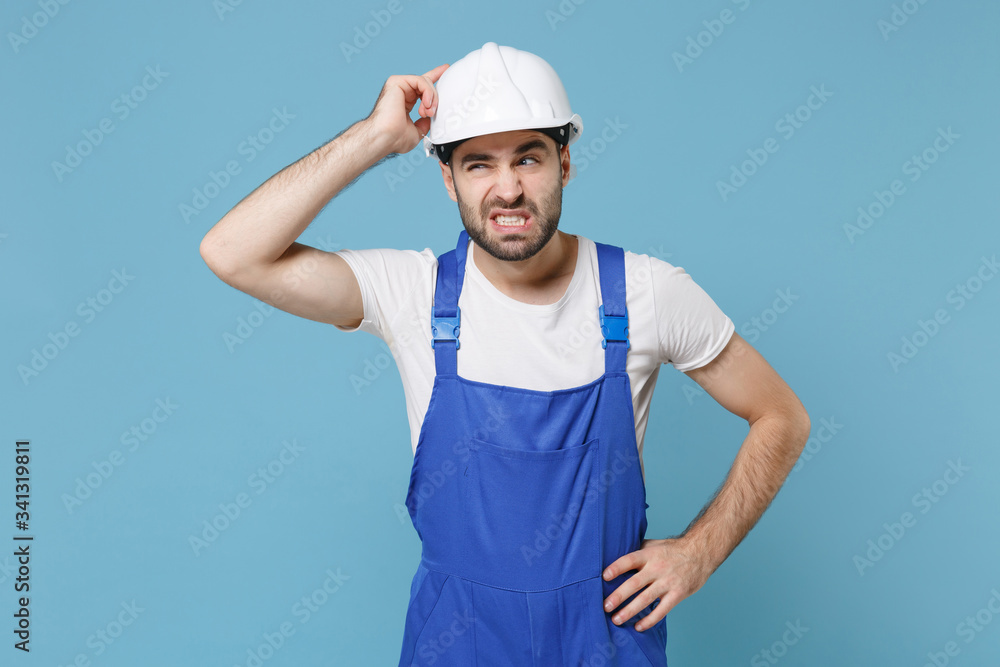 Preoccupied young man in coveralls protective helmet hardhat isolated on blue background. Instruments accessories for renovation apartment room. Repair home concept. Put hand on head, looking aside.