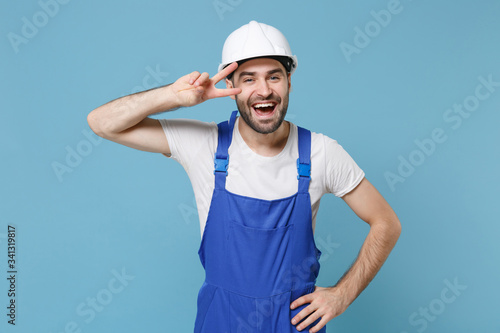 Laughing young man in coveralls protective helmet hardhat isolated on blue wall background in studio. Instruments accessories for renovation apartment room. Repair home concept. Showing victory sign.