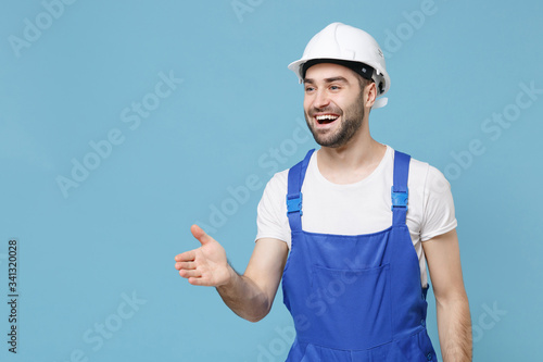 Cheerful man in coveralls protective helmet hardhat isolated on blue background. Instruments accessories for renovation apartment room. Repair home concept. Stand with outstretched hand for greeting.