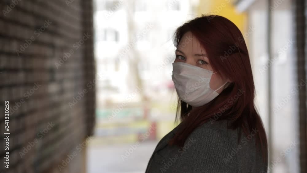 A beautiful woman in a coat, with red hair, European appearance turns and looks at the camera on a city street. The camera follows from behind. A girl in a medical mask. pandemic. Coronavirus A beautiful woman in a coat, with red hair, European appearance turns and looks at the camera on a city street. The camera follows from behind. A girl in a medical mask. pandemic. Coronavirus