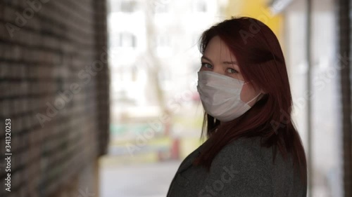 A beautiful woman in a coat, with red hair, European appearance turns and looks at the camera on a city street. The camera follows from behind. A girl in a medical mask. pandemic. Coronavirus photo