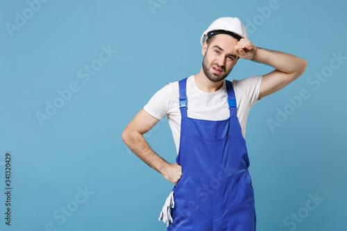 Exhausted tired young man in coveralls protective helmet hardhat isolated on blue wall background studio. Instruments accessories for renovation apartment room. Repair home concept. Put hand on head.