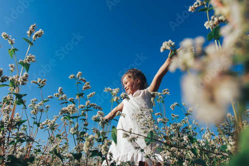 Cheerful little girl in white linen dress standing with open arms on flowery meadow or field. Cute child playing among spring flowers outdoors. Summer, nature, childhood concept