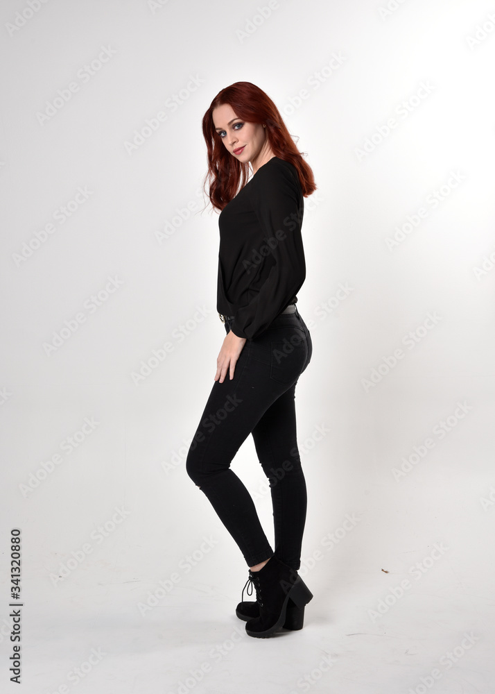 Portrait of a pretty girl with red hair wearing black jeans, boots and a blouse. full length standing pose in side profile, on a studio background. Portrait of a pretty girl with red hair wearing black jeans, boots and a blouse. full length standing pose in side profile, on a studio background.
