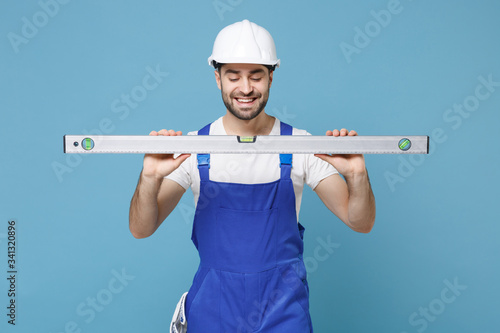 Smiling young man in coveralls protective helmet hardhat hold building bubble spirit level isolated on blue wall background. Instruments accessories for renovation apartment room. Repair home concept.