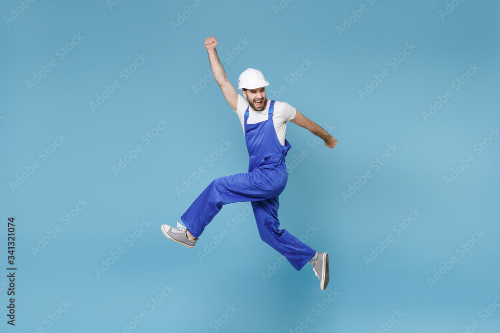 Overjoyed young man in coveralls protective helmet hardhat isolated on blue background. Instruments accessories renovation apartment room. Repair home concept. Jumping clenching fists like winner. Overjoyed young man in coveralls protective helmet hardhat isolated on blue background. Instruments accessories renovation apartment room. Repair home concept. Jumping clenching fists like winner.