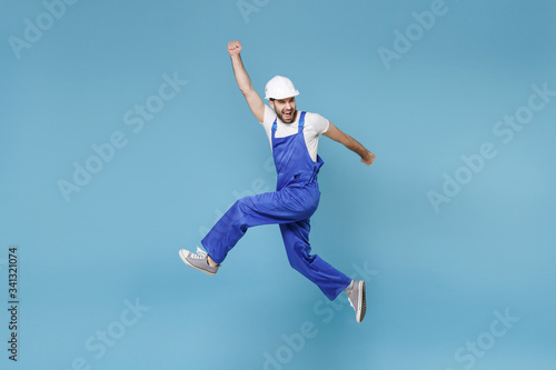 Overjoyed young man in coveralls protective helmet hardhat isolated on blue background. Instruments accessories renovation apartment room. Repair home concept. Jumping clenching fists like winner.