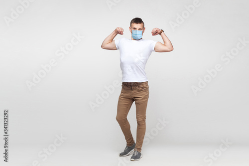 I am strong. Full length portrait of young man in white shirt with surgical medical mask standing and showing his biceps and looking with haughty face. indoor studio shot, isolated on gray background.
