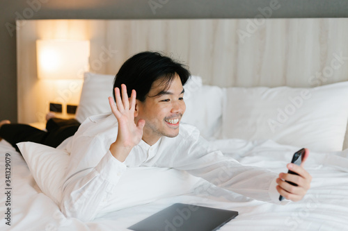 Asian male in white shirt using mobile phone while working on bed. work from home concept.