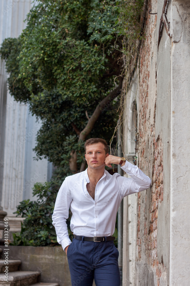 Fototapeta premium handsome and young guy in a white shirt enjoy the summer weather leans on a brick wall in Venice.