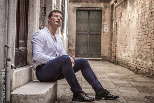 young man sits on the steps in a white shirt and classic black shoes in venice on the street