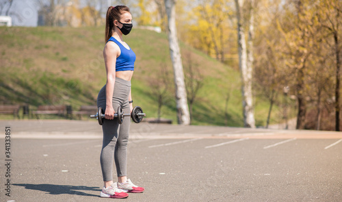 Young athletic girl doing exercises with dumbbells in a medical mask on the playground during a pandemic. Squats COVID-19. Health care.