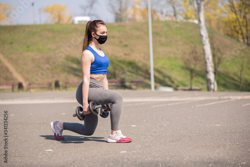 Athletic beautiful girl in a medical mask lunges with dumbbells on the playground during a pandemic. COVID-19. Health care.