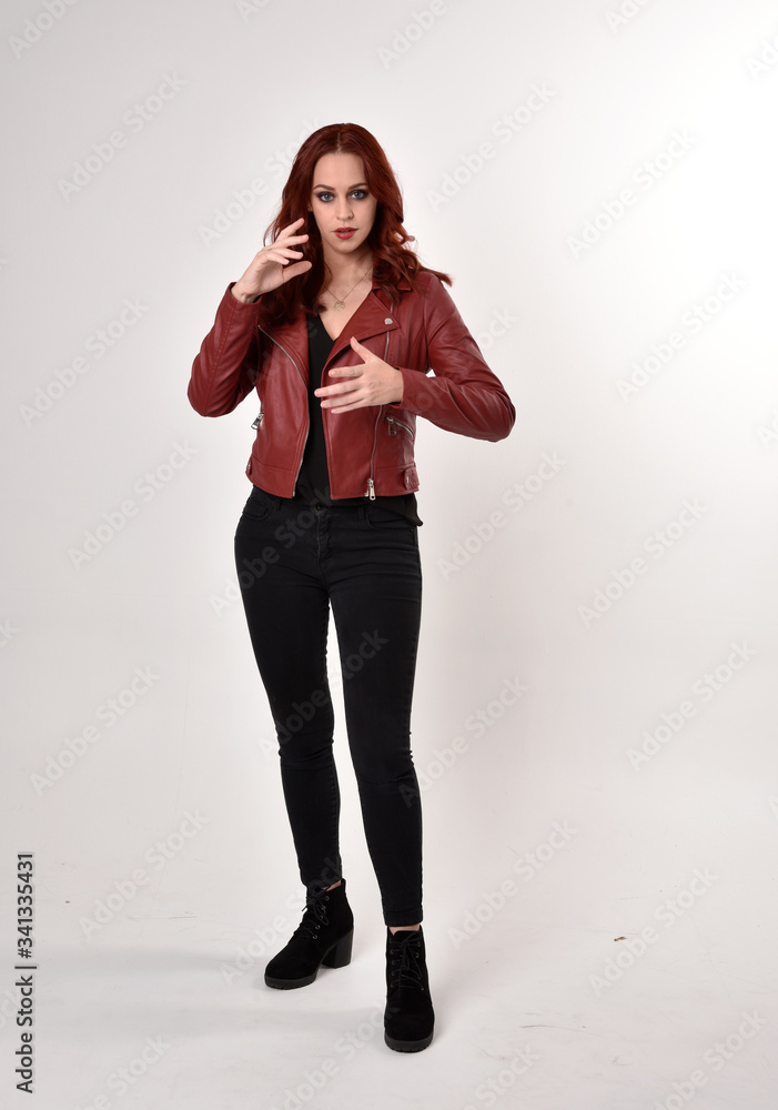 Portrait of a pretty girl with red hair wearing black jeans and boots with leather jacket. full length standing pose with hand gesture on a studio background. Portrait of a pretty girl with red hair wearing black jeans and boots with leather jacket. full length standing pose with hand gesture on a studio background.