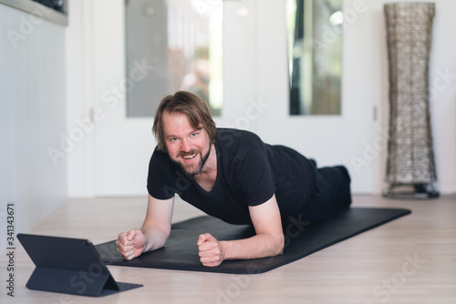 Mann macht Planks zu Hause auf der Fitnessmatte mittels Online-Kurs und lächelt in die Kamera photo
