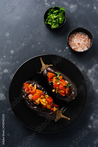Baked stuffed eggplant with different vegetables, tomato, pepper, onion and parsley on a black stone or concrete table background. Top view.